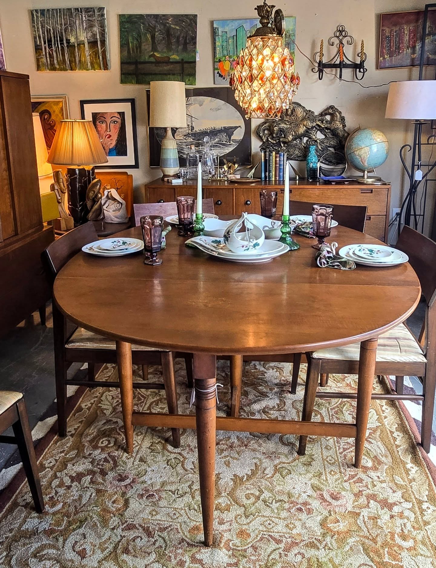 Round wooden dining table set with plates, glasses, and cutlery, surrounded by chairs, in a room with artwork and lamps.