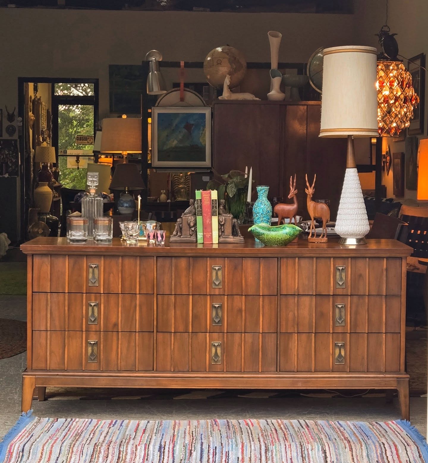 Wooden sideboard with decorative items, lamps, and books, placed on a colorful striped rug in a room with various furniture and decor.