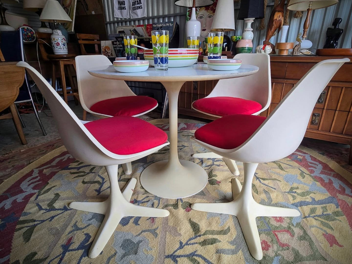 Round table with four white chairs with red cushions, set with colorful cups and plates, in a room with wooden furniture.