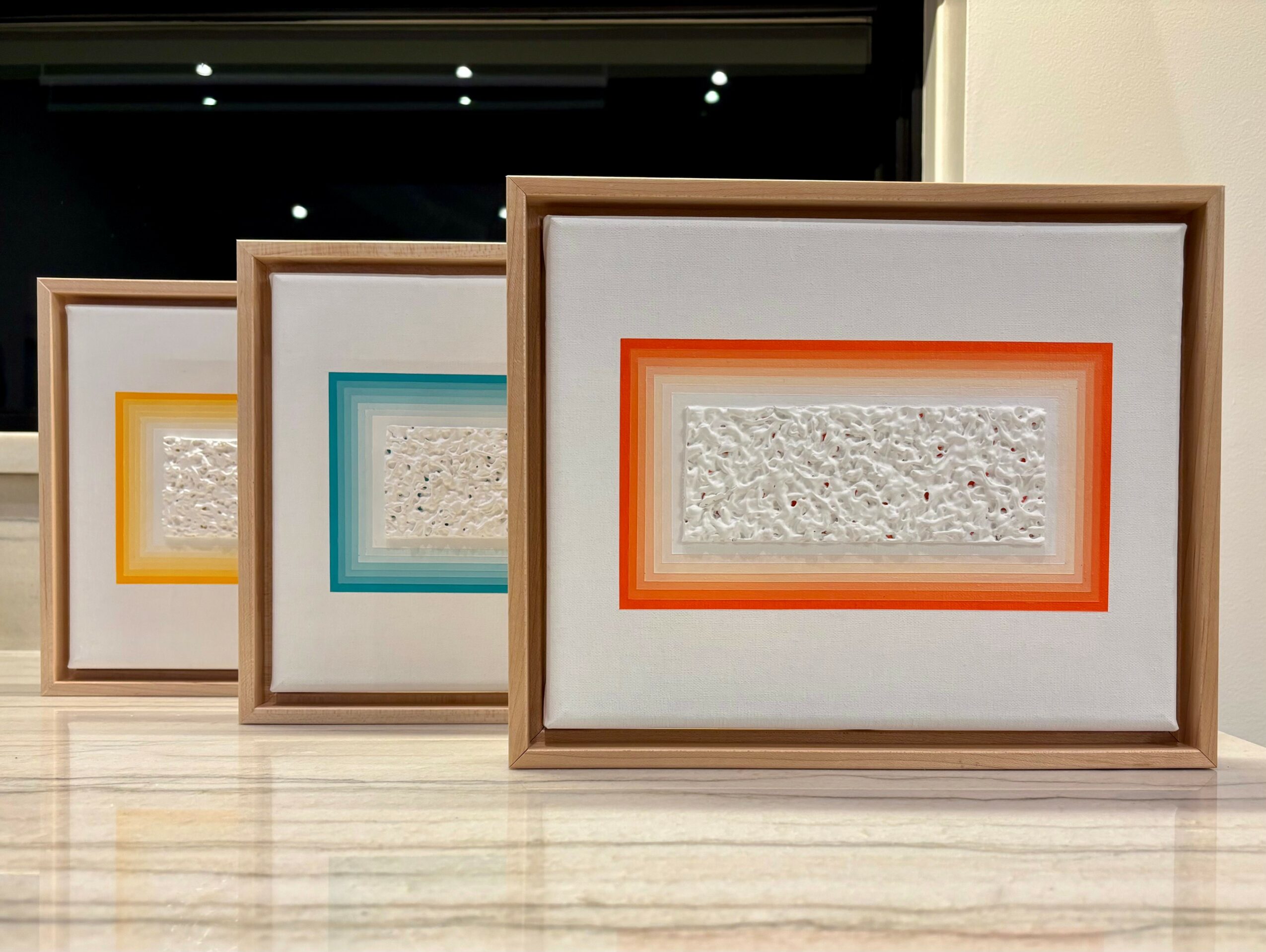 Three framed textured art pieces with colorful borders, arranged on a light-colored surface, with a dark background behind them.