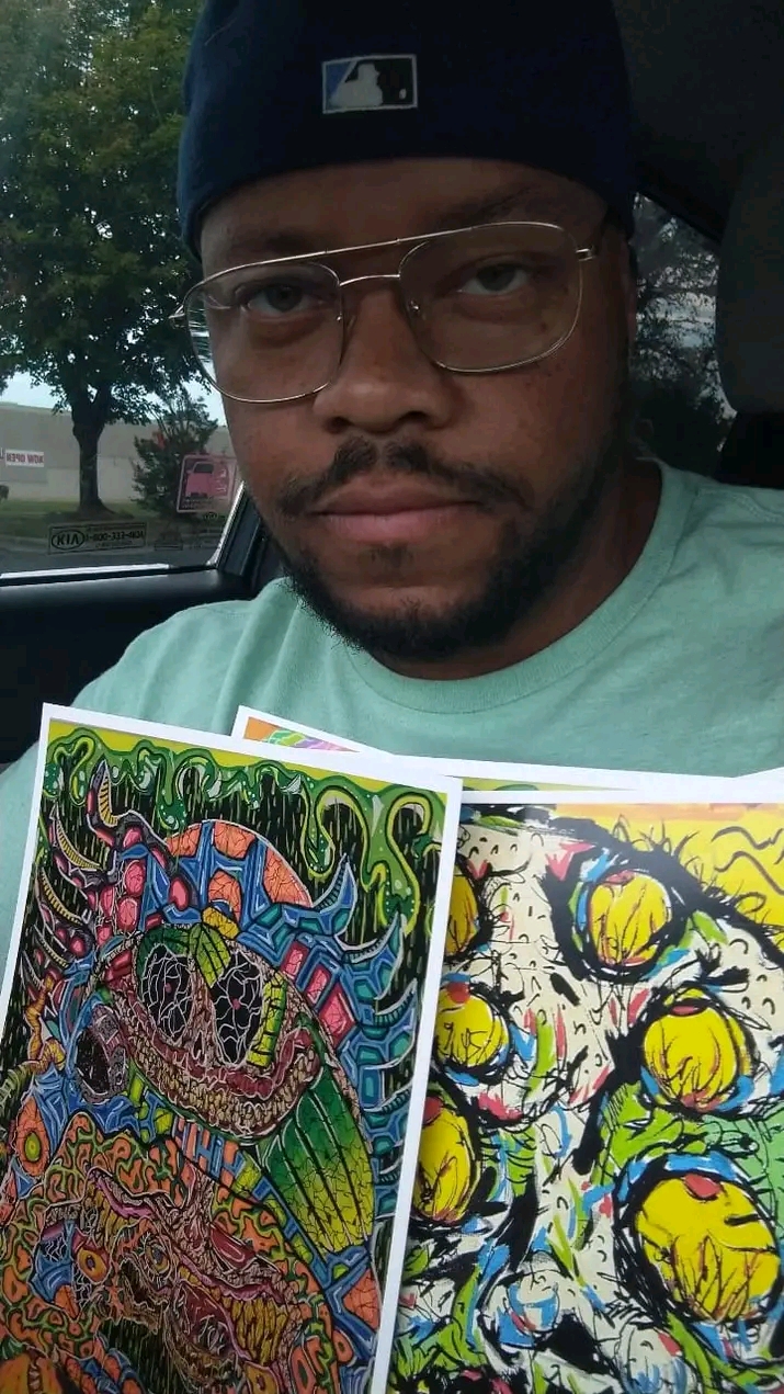 Person wearing glasses and a cap holding two colorful abstract art pieces, outdoors with trees in background.