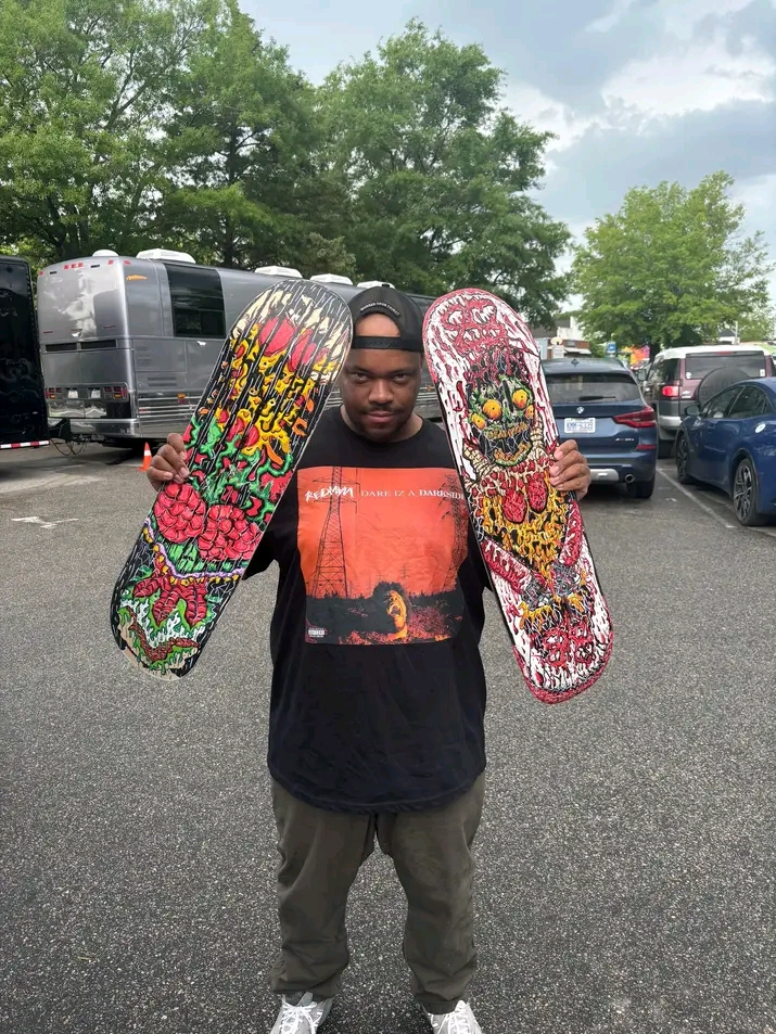 Young man holding two skateboard decks with colorful, floral, and graphic designs in a parking lot with trees and vehicles.