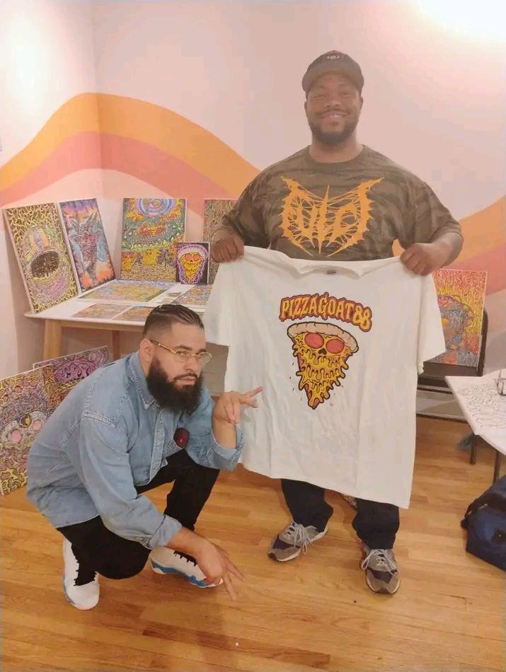 Two men pose indoors, one holding a white T-shirt with a pizza graphic, colorful artwork displayed behind them.