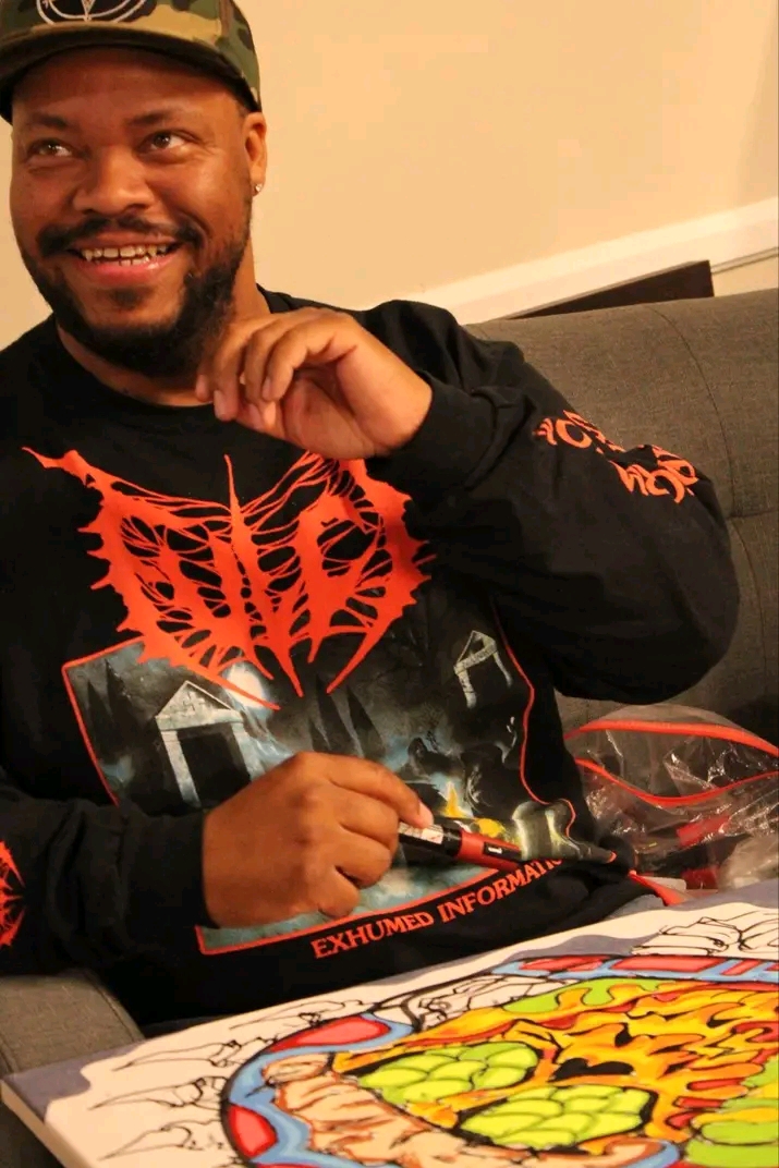 Person with a beard smiling, wearing a black cap and a black sweatshirt with red and orange designs, sitting at a table with colorful artwork.