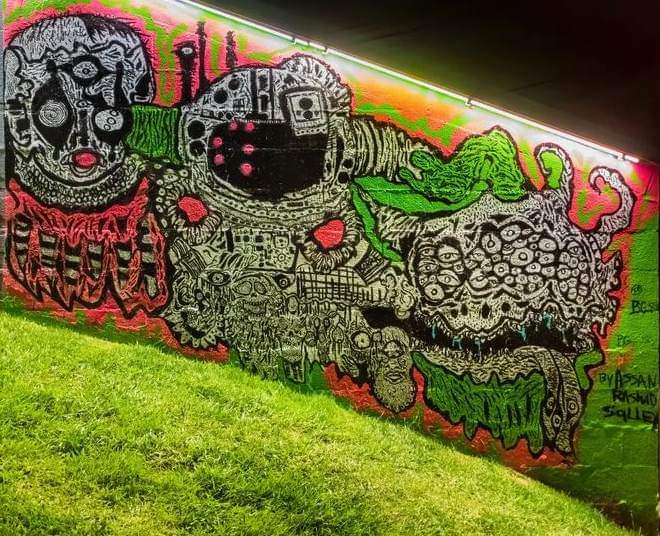 Colorful graffiti mural with abstract faces, shapes, and patterns on a wall, with grass in the foreground.