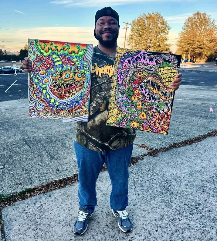 Person holding two colorful abstract paintings outdoors, smiling, with trees and parking lot in background.
