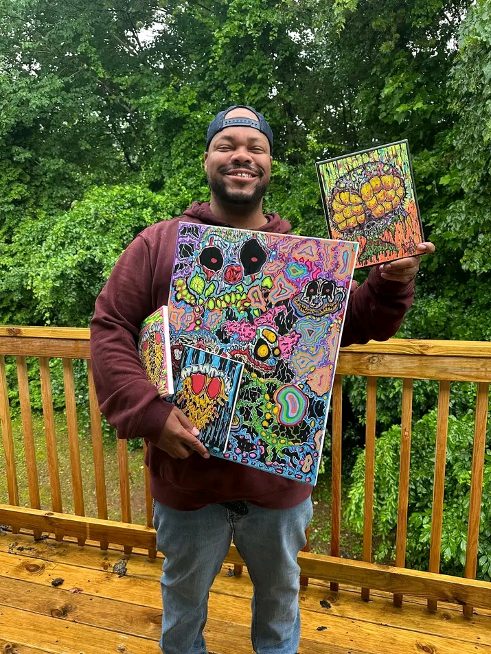 Smiling man holding colorful paintings outdoors on a wooden deck with green trees in background.