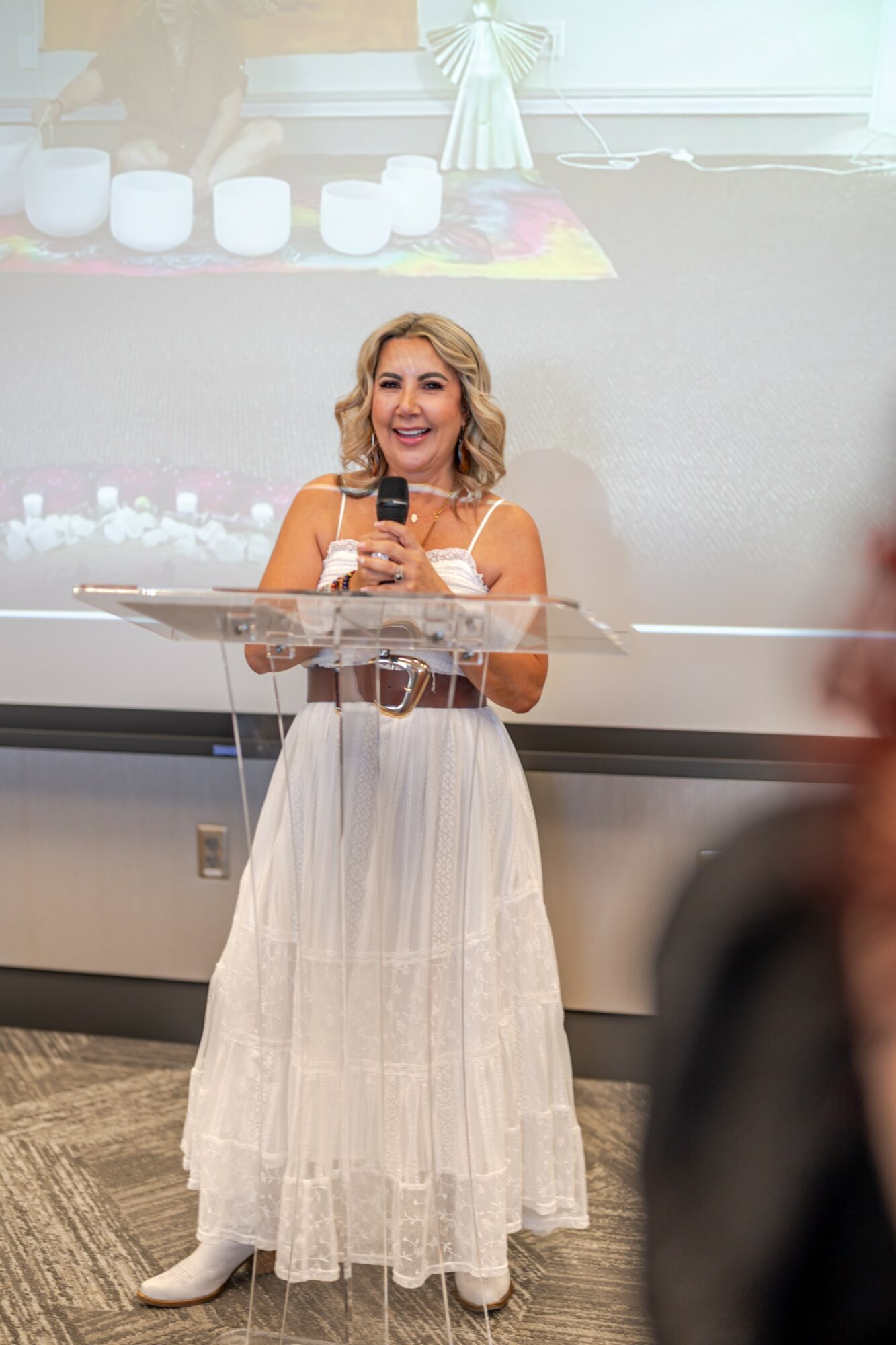 Woman with blonde hair in a white dress holding a microphone, smiling, standing behind a clear podium in a room.