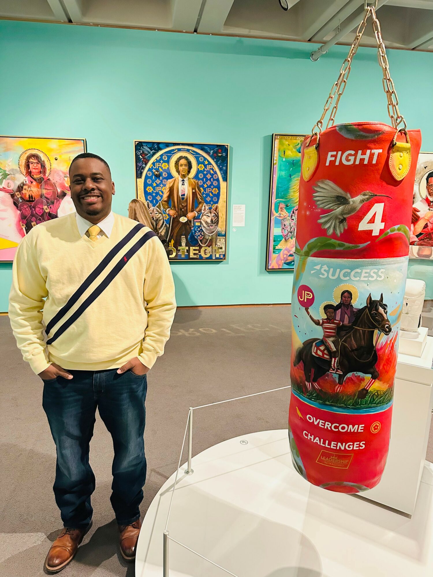 Man standing next to a colorful punching bag with motivational words, in an art gallery with paintings on the wall.