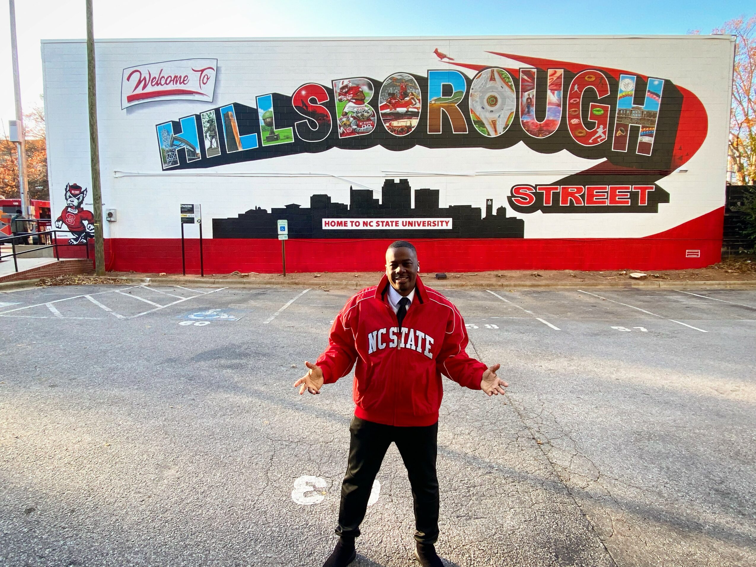 Person standing in front of a mural with colorful text reading 'Hillsborough' and a cityscape silhouette, wearing a red NC State sweatshirt.
