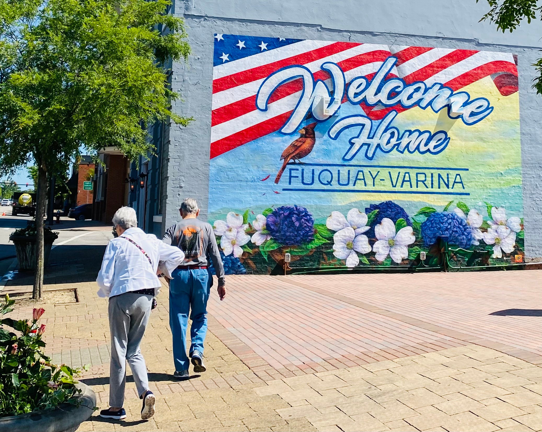 People walking past a colorful mural with a welcome message, American flag design, and flowers, on a sunny day.
