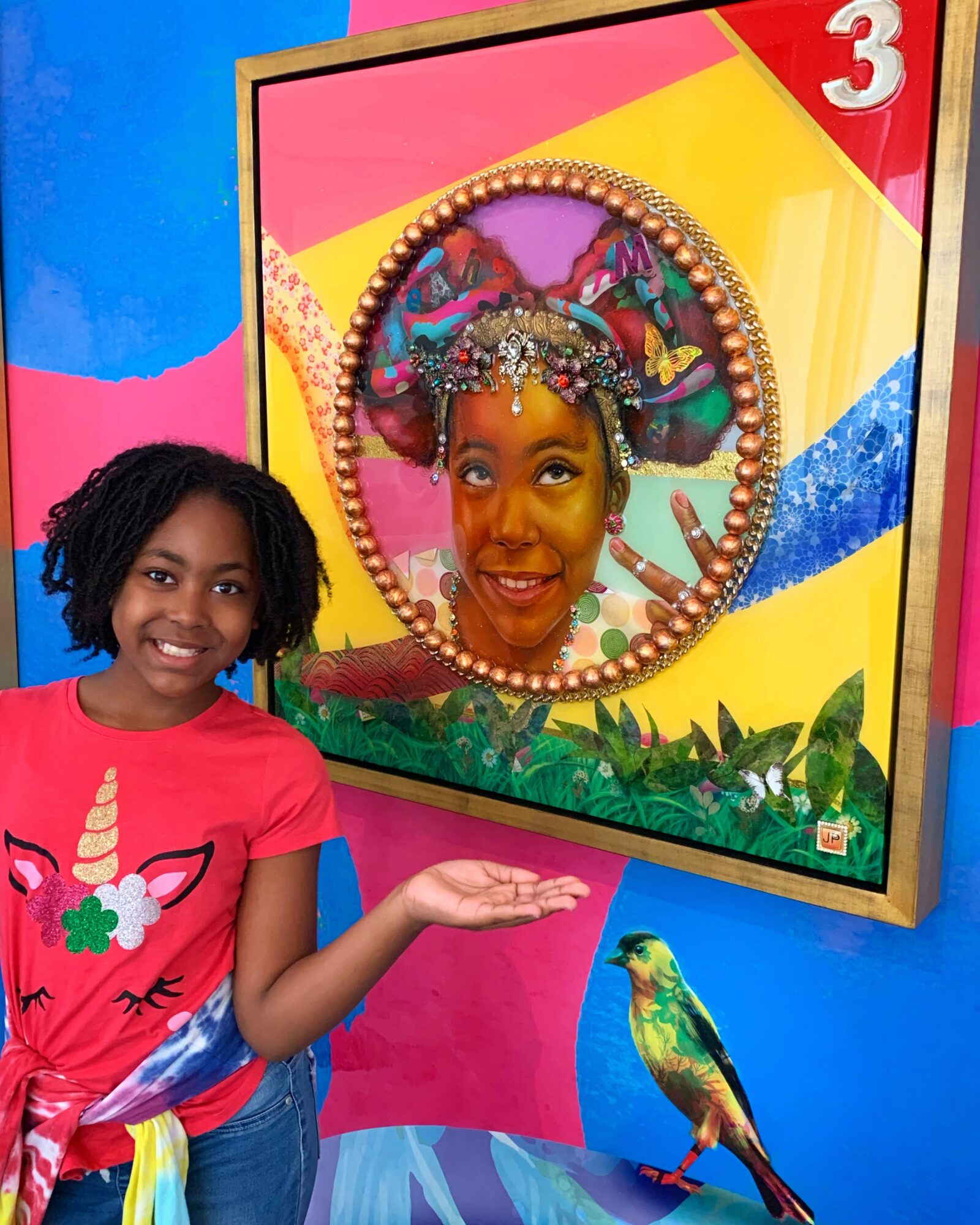 Child smiling and pointing at a colorful mirror with a person’s face reflected, decorated with a headpiece, in a vibrant setting.