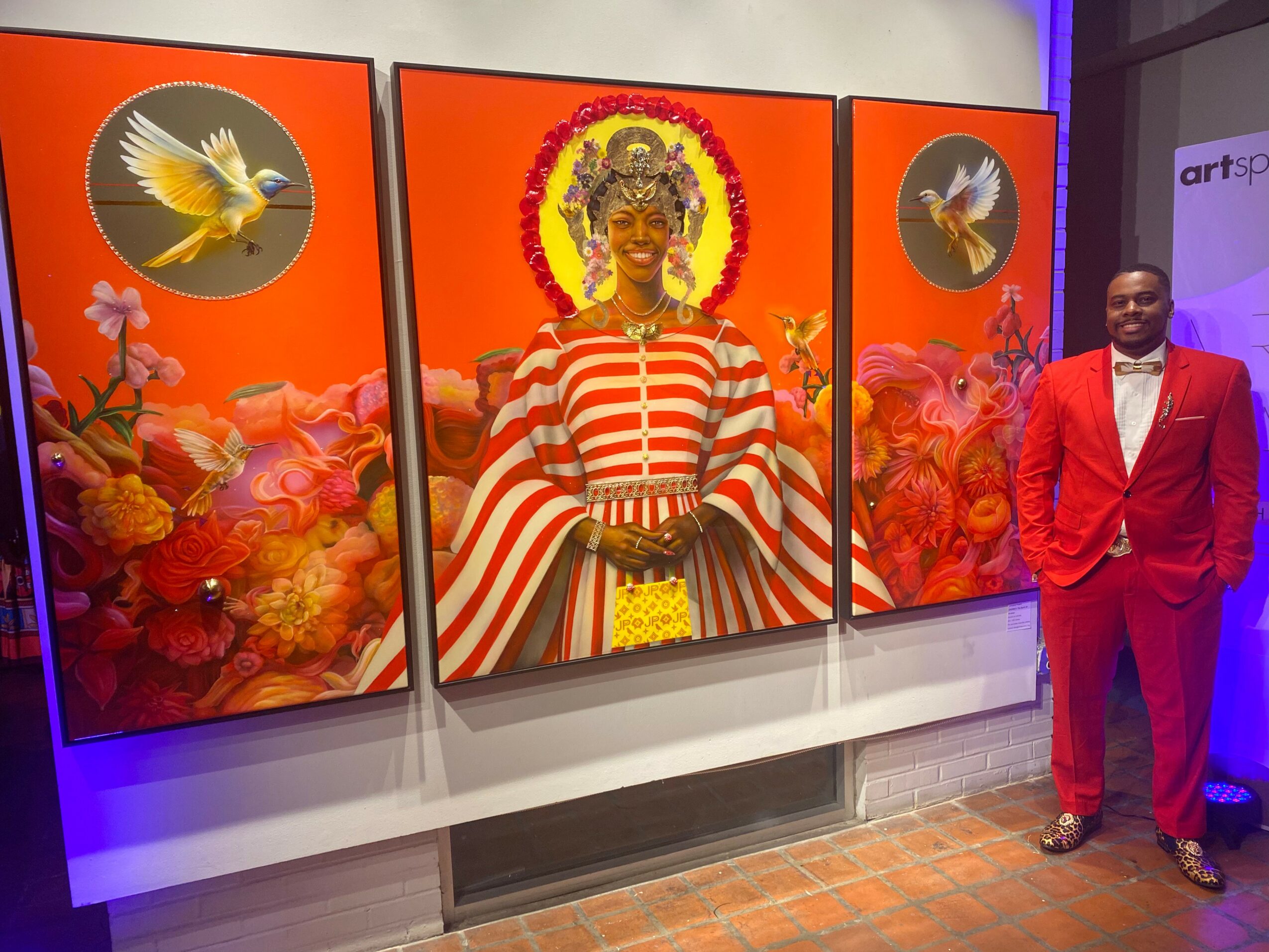 Man in red suit standing next to colorful artwork featuring a central figure with a crown, surrounded by flowers and birds.