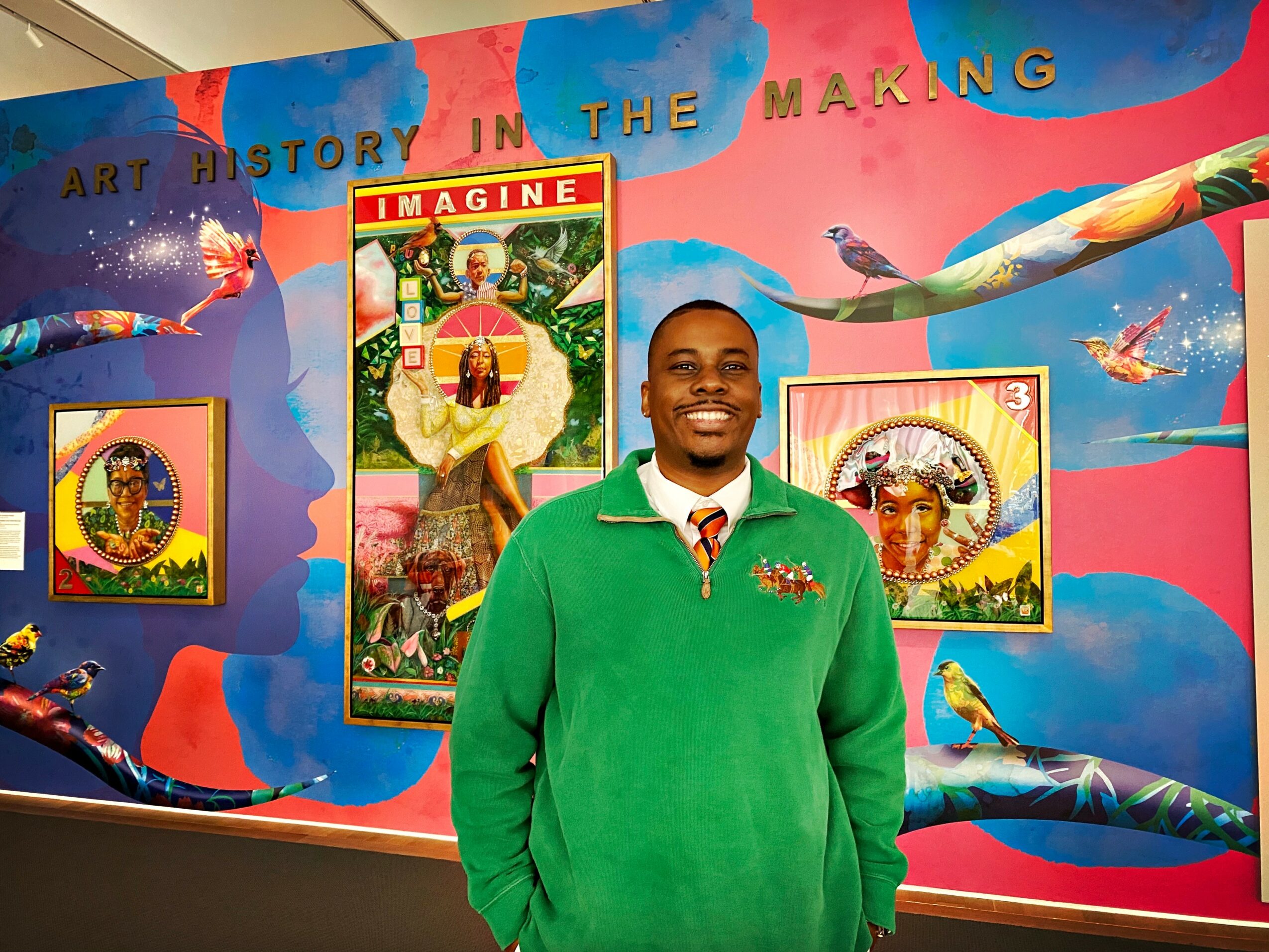 Young man in green sweater smiling in front of colorful art display with paintings and a pink and blue background.