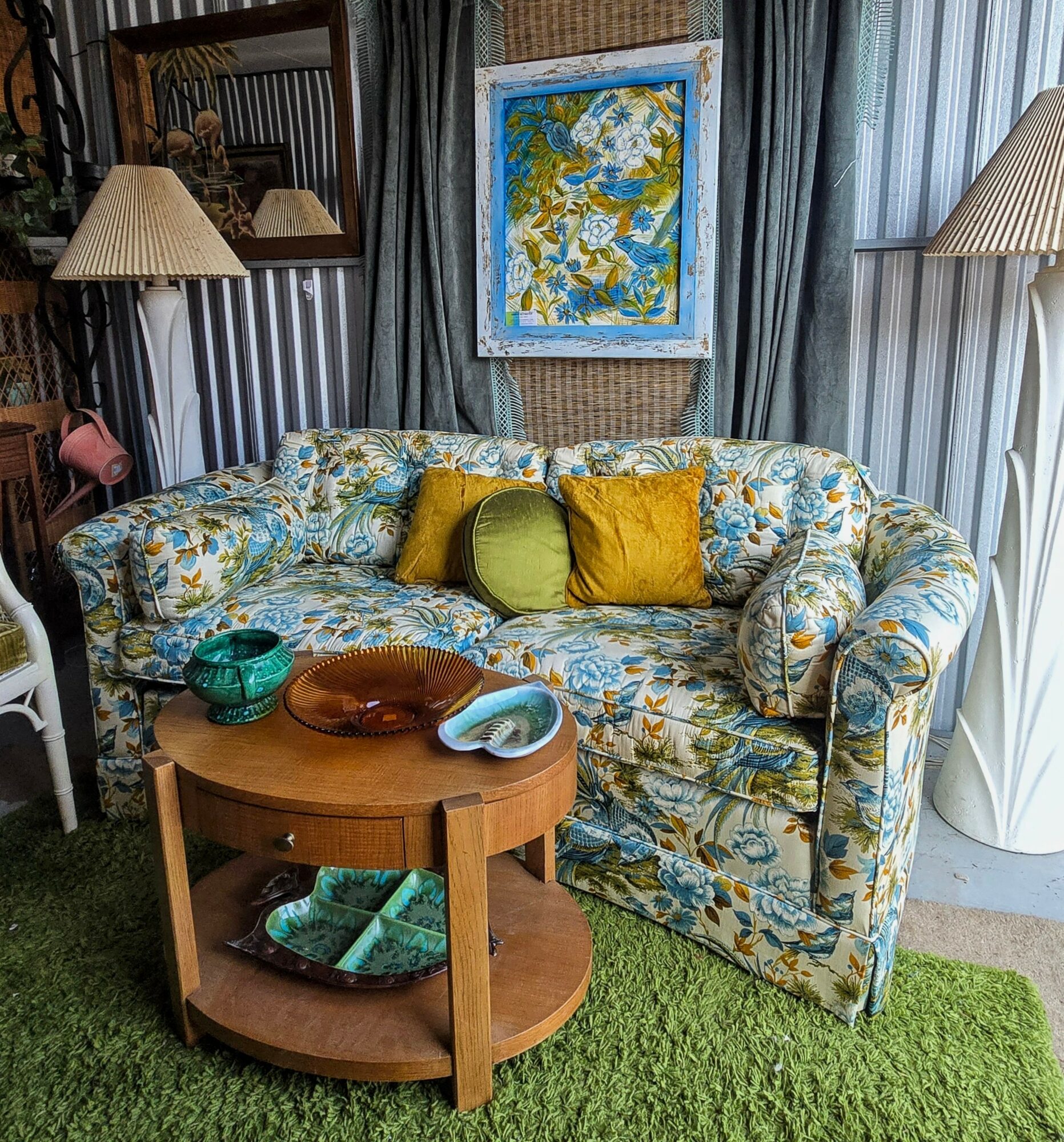 Living room with floral sofa, wooden table, lamps, and framed artwork on curtains, green rug on floor.