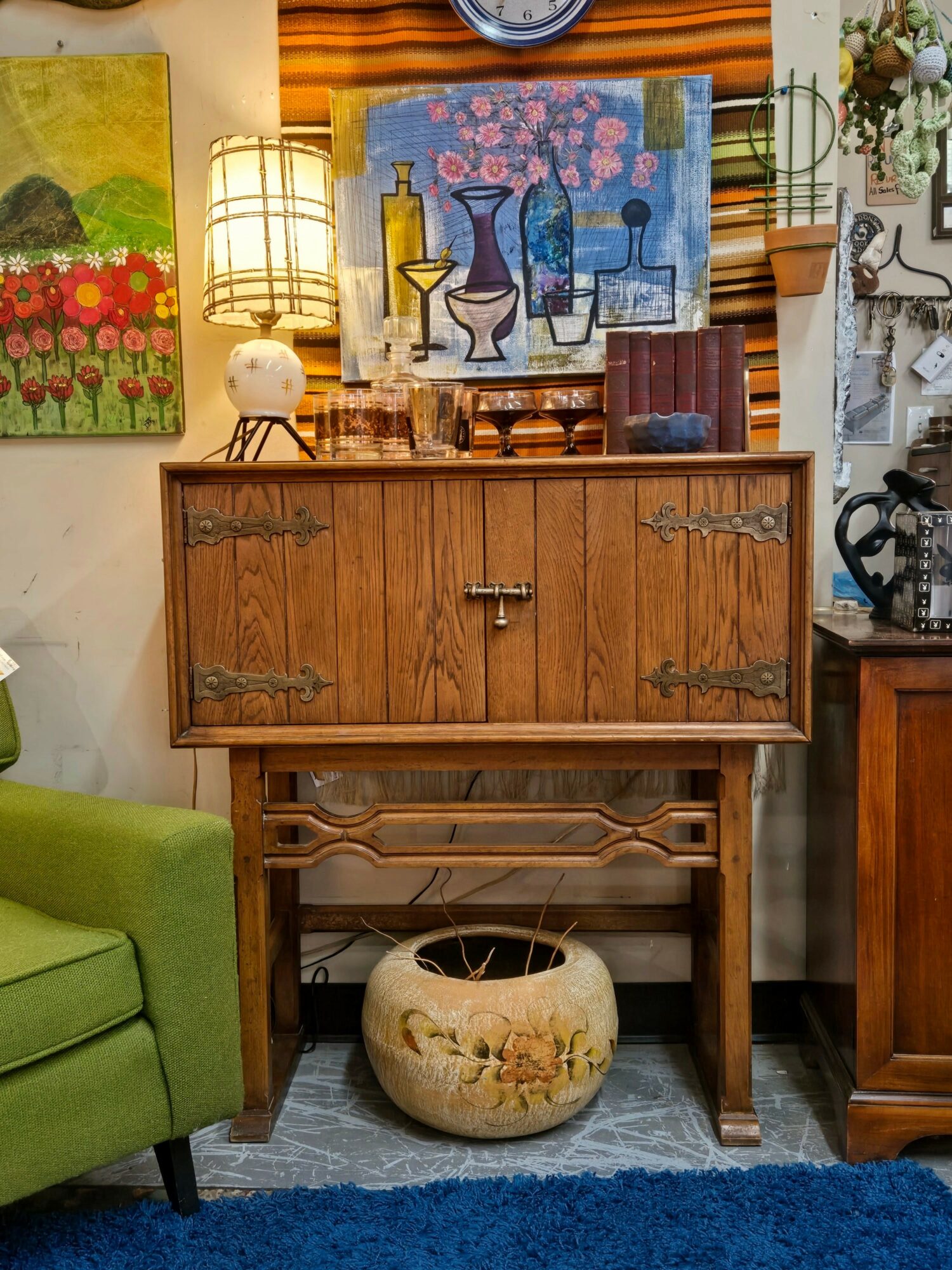 Wooden cabinet with artwork on top, a large ceramic pot underneath, and surrounding furniture and decorations.