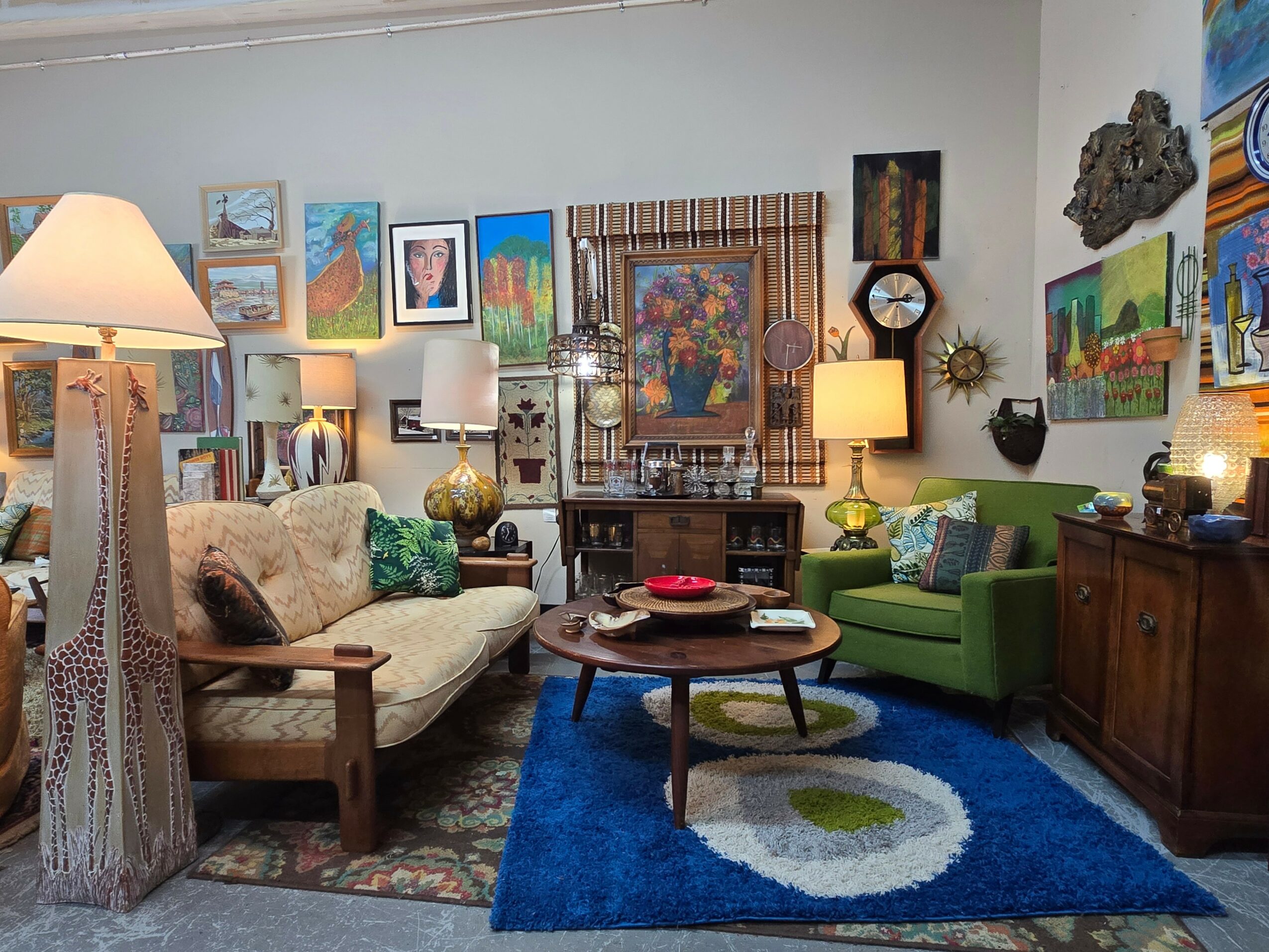 Living room with colorful paintings, a beige sofa, green armchair, wooden coffee table, and blue rug, illuminated by lamps.