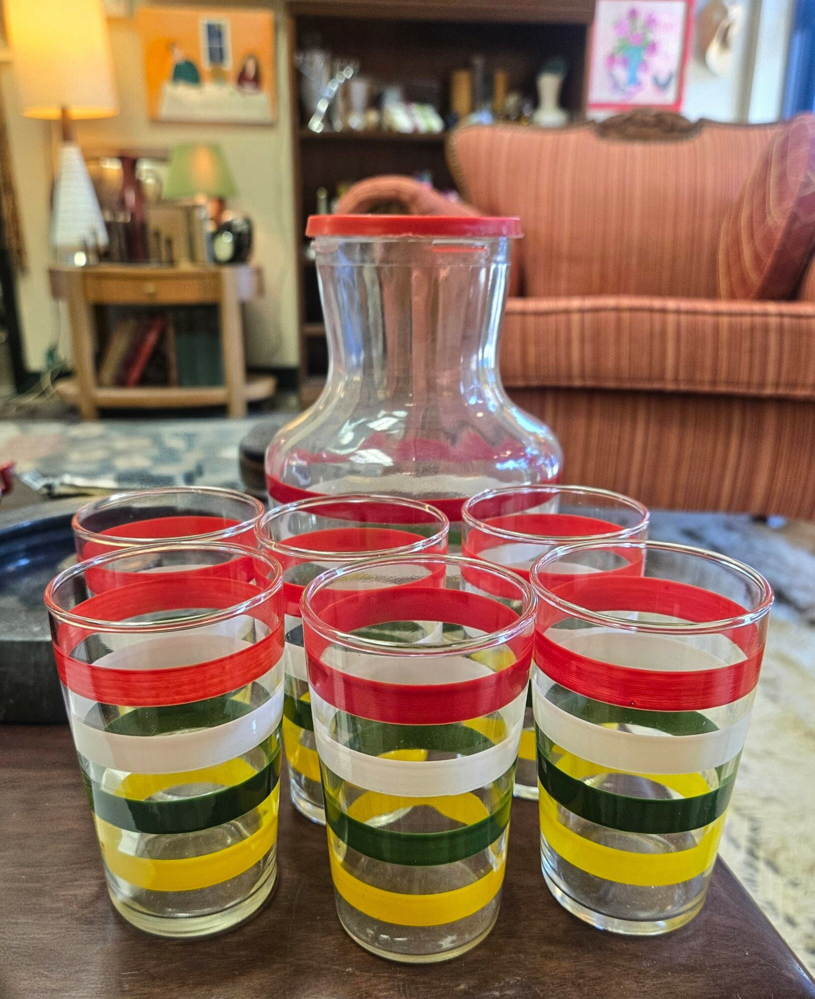 A clear glass pitcher surrounded by seven matching glasses with colorful horizontal stripes, on a wooden table in a living room.