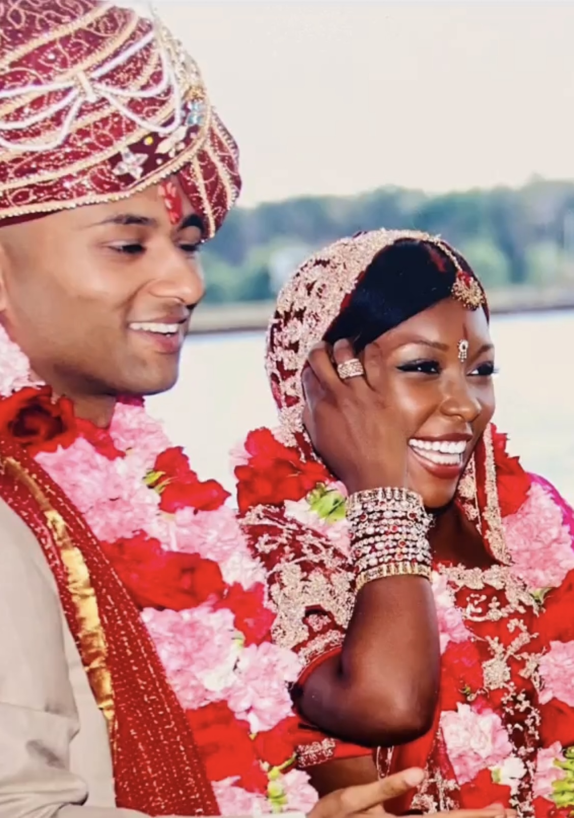 Smiling couple dressed in traditional wedding attire, adorned with floral garlands and jewelry, outdoors with a blurred background.