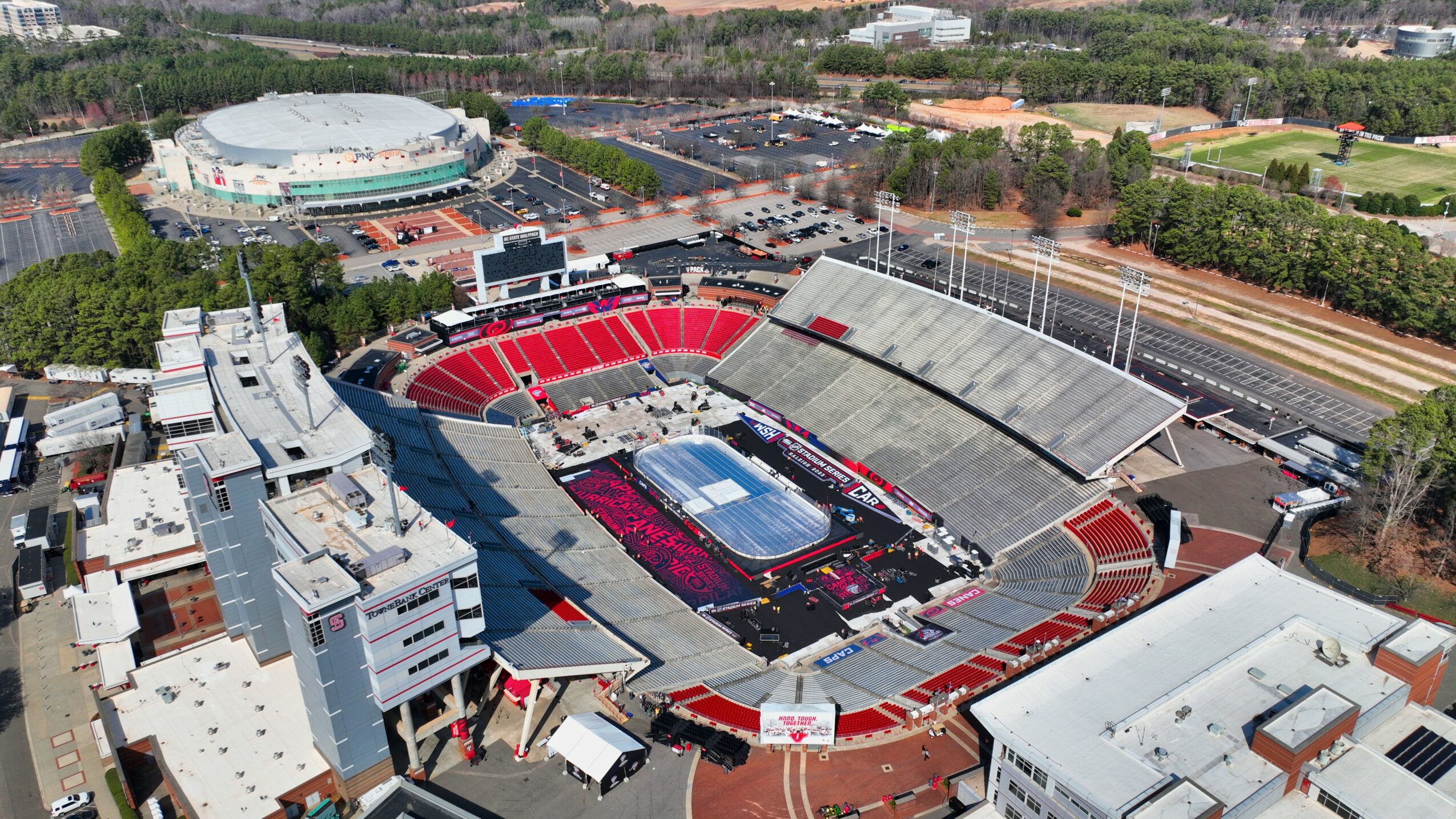 Aerial view of a stadium with red seating, a central field, and surrounding buildings, with trees and roads nearby.