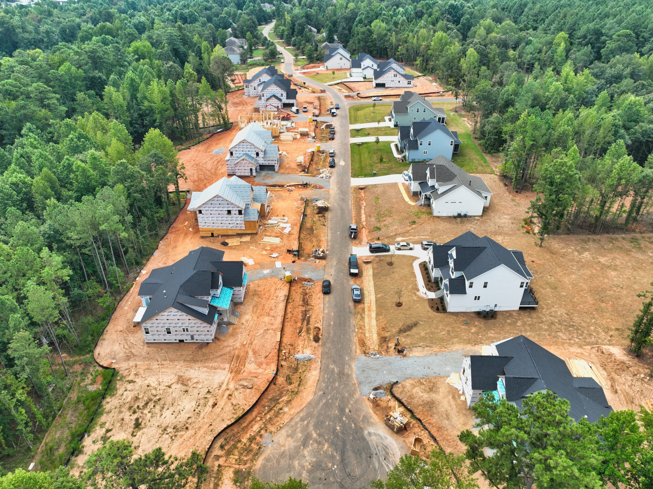 Aerial view of a residential neighborhood with houses under construction, surrounded by trees and a dirt road in the center.