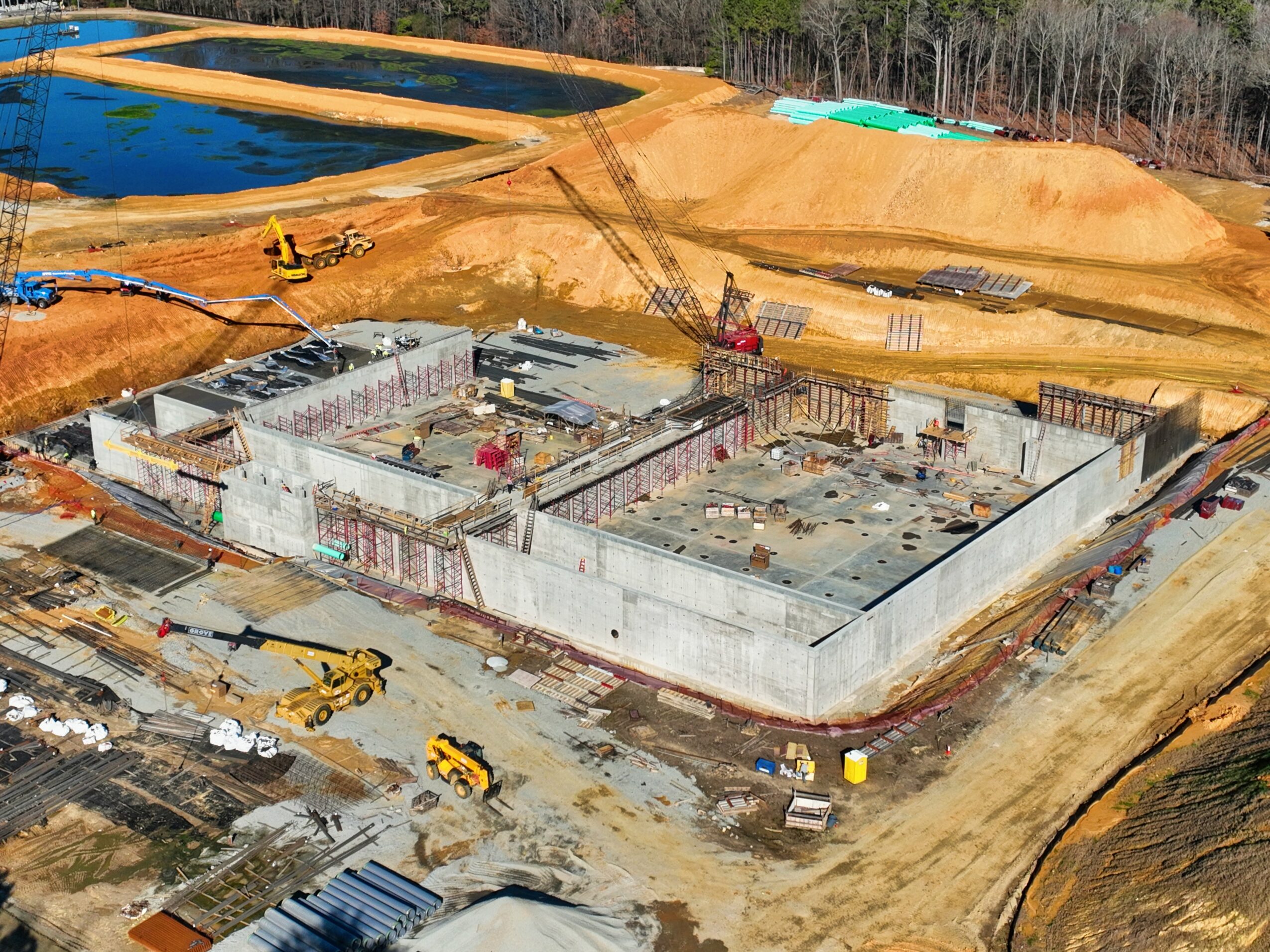 Construction site with a large concrete foundation, surrounded by dirt and construction equipment, with a body of water nearby.