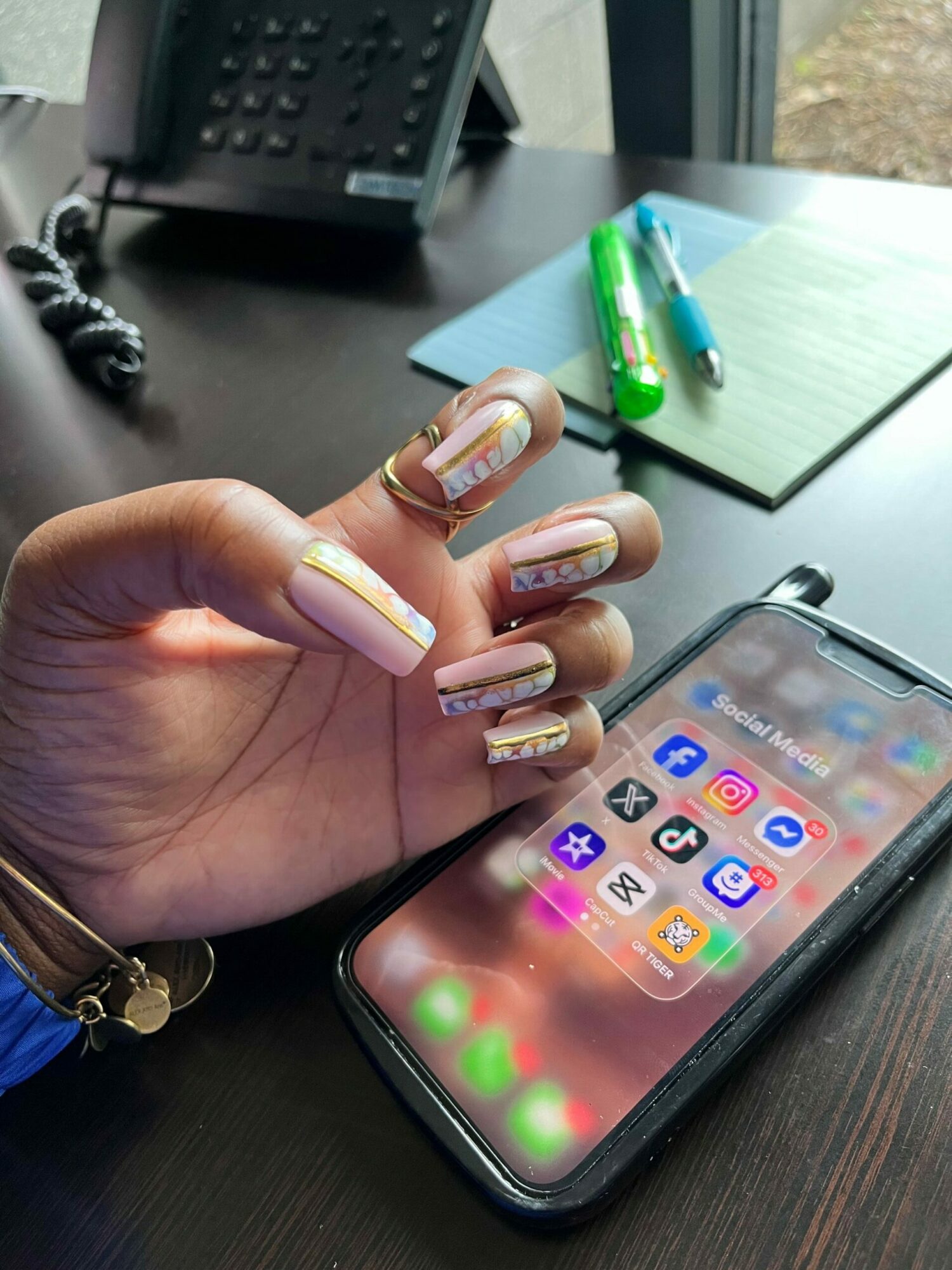 Hand with decorated nails near smartphone on dark wooden surface, with office supplies in background.