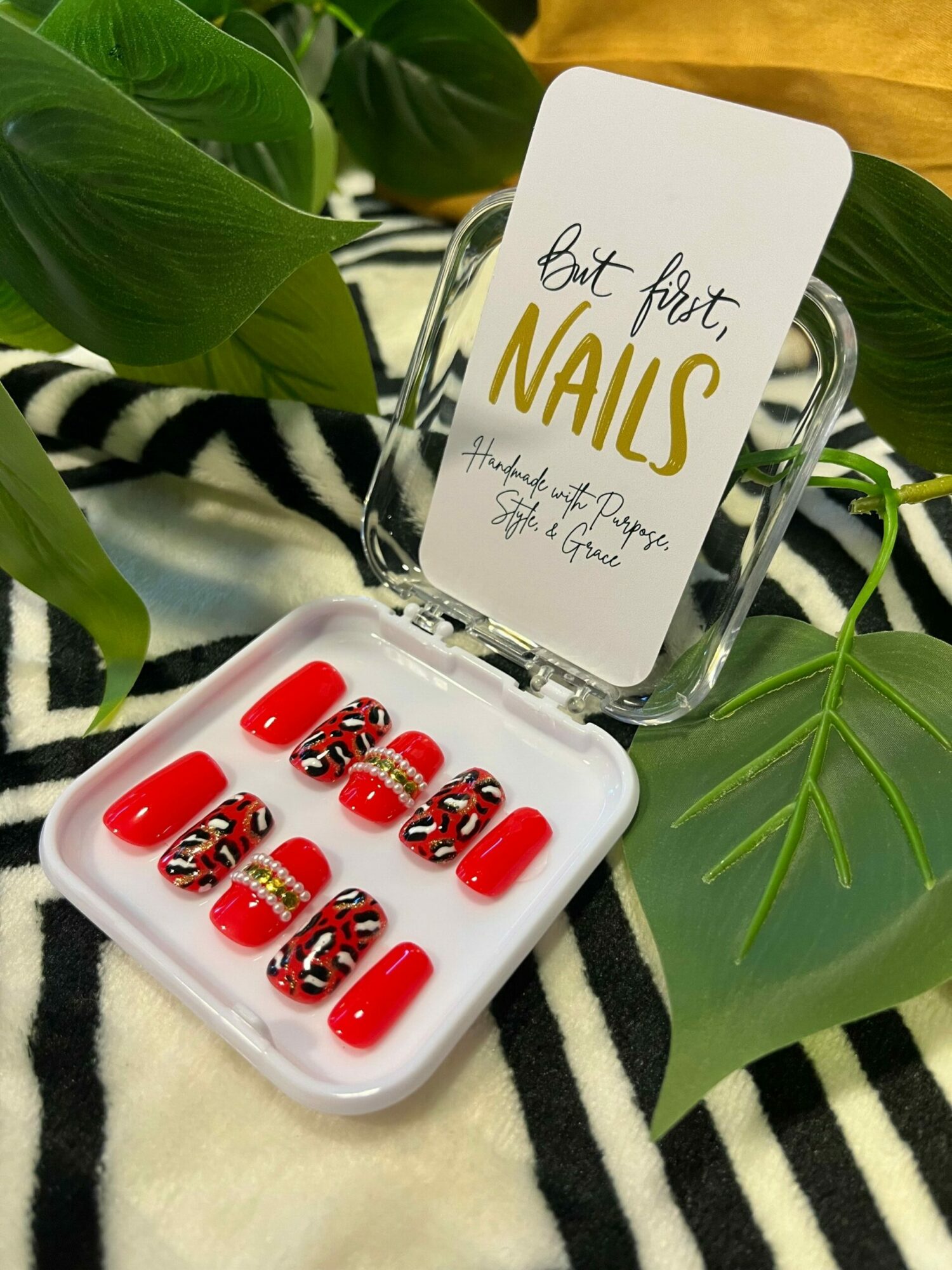 Nail polish set with red and decorated nails, placed on a black and white striped fabric with green leaves nearby.