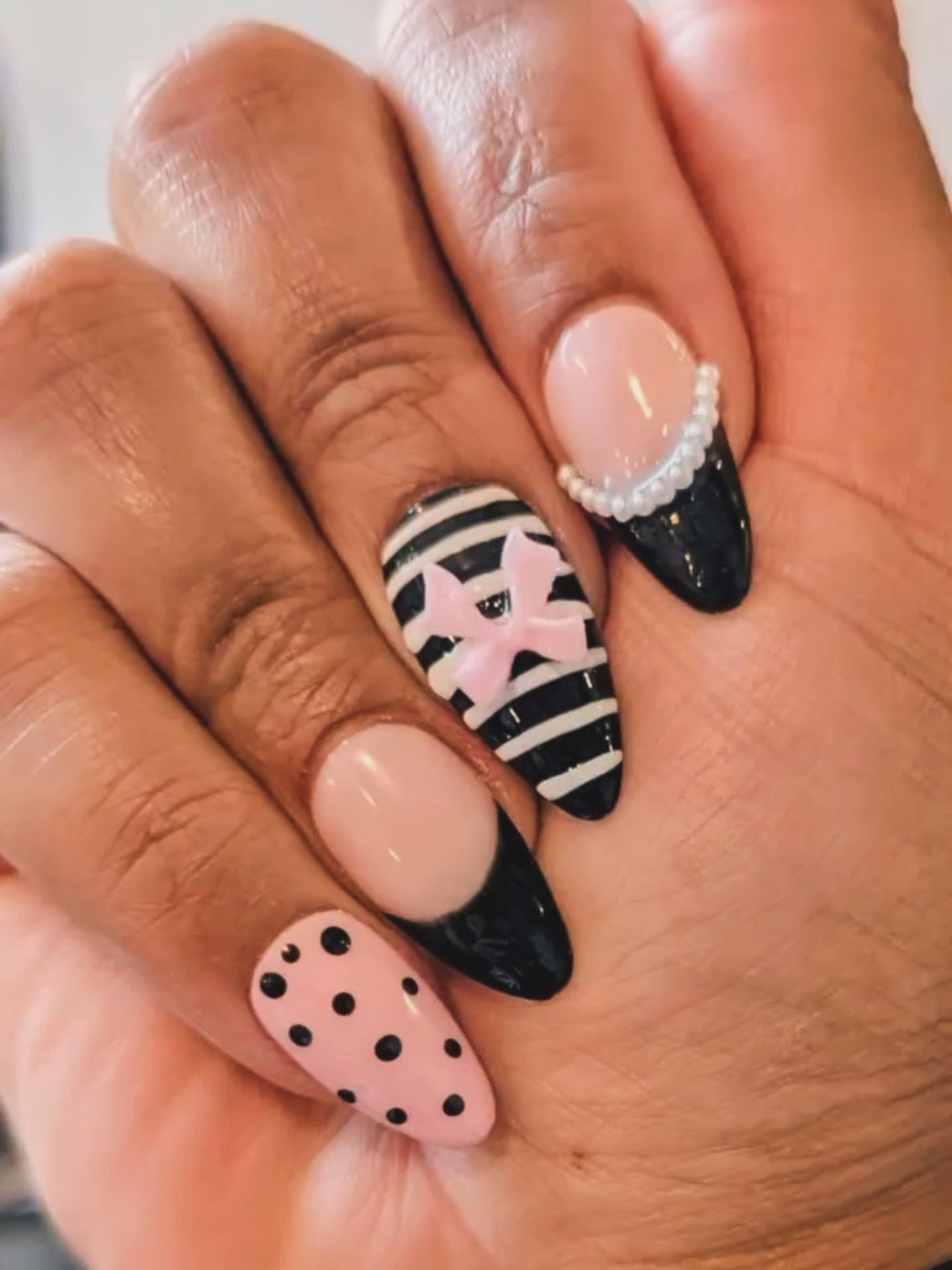 Close-up of four fingers with decorated nails, featuring black, pink, and striped designs, and small pearl accents.