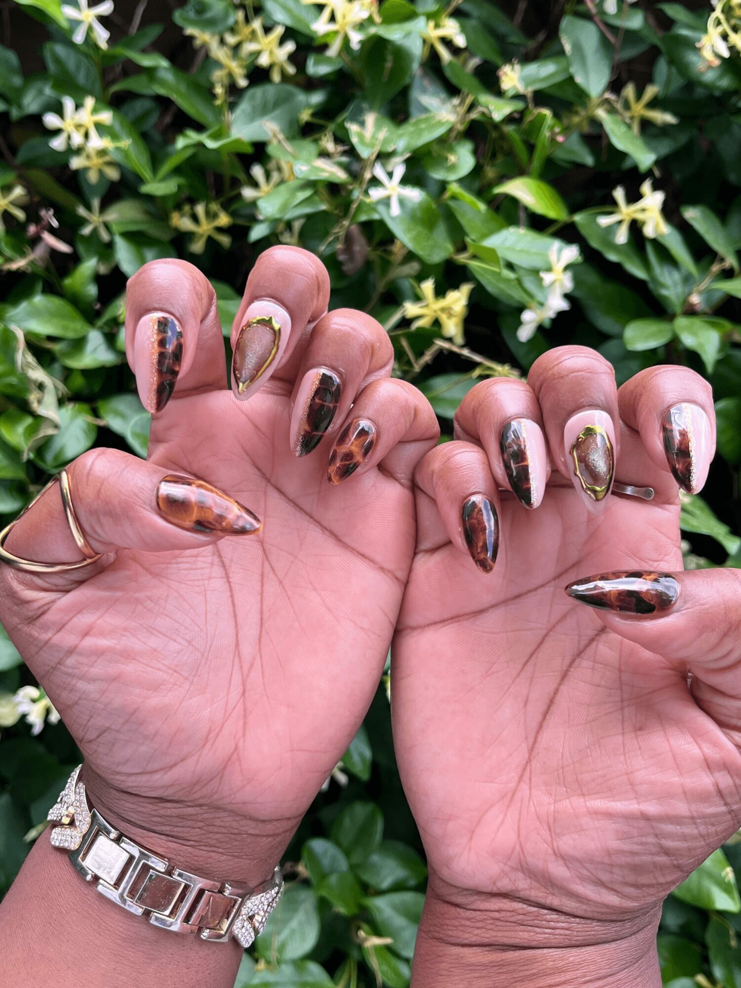 Hands with decorated nails held against green leafy background with small white flowers, wearing a silver watch and rings.