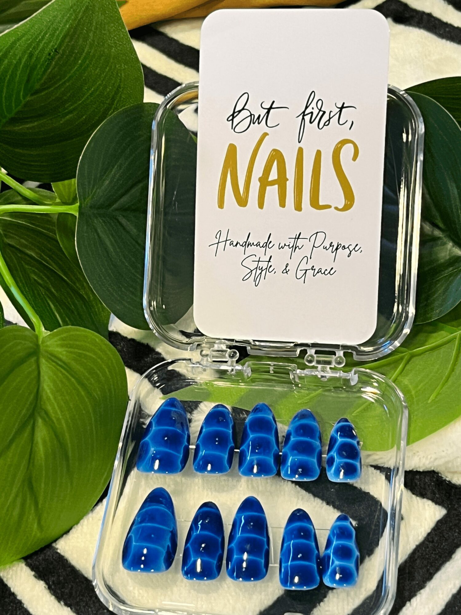 Open clear case with blue press-on nails inside, surrounded by green leaves and a black-and-white striped fabric background.