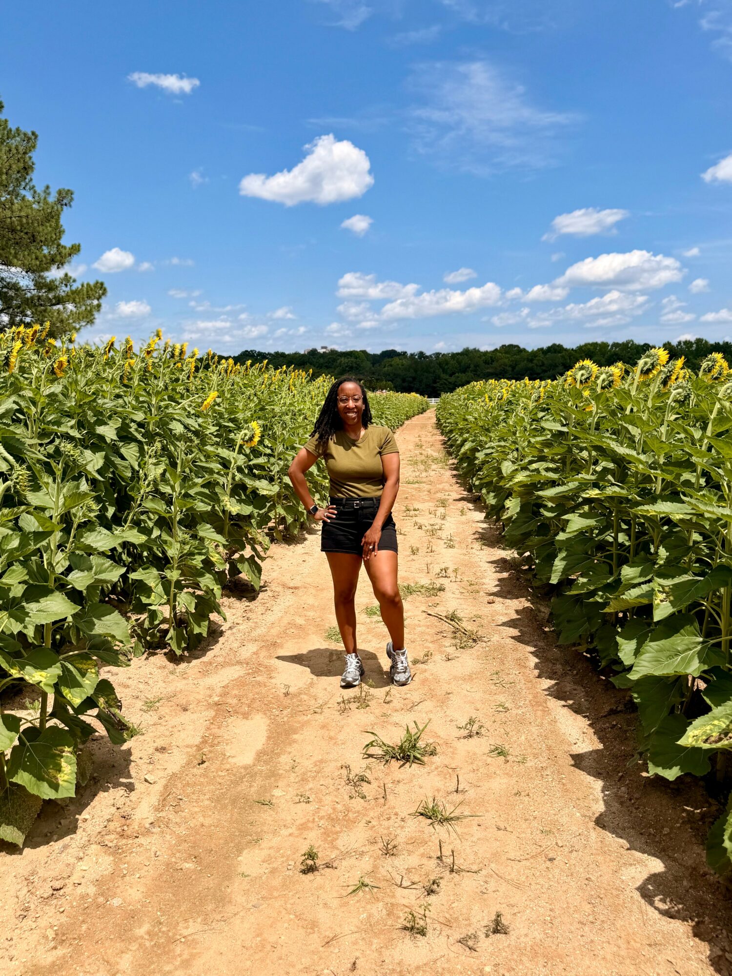 Woman standing on a dirt path between sunflower fields under a blue sky with clouds.