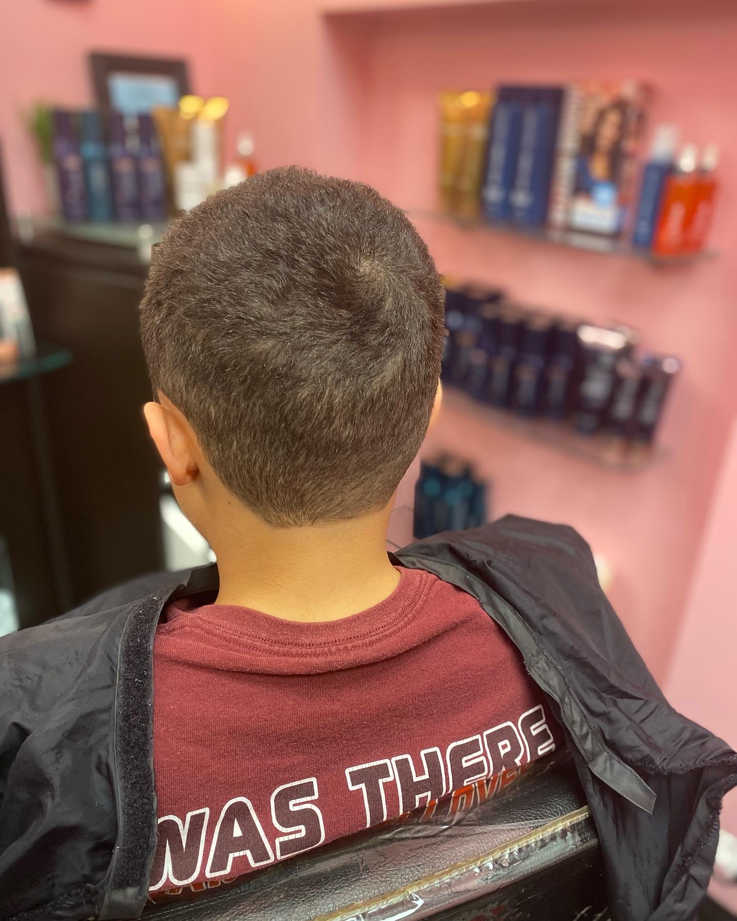 Back of a person's head with short hair, wearing a maroon shirt and black jacket, in a store with pink walls and shelves of products.