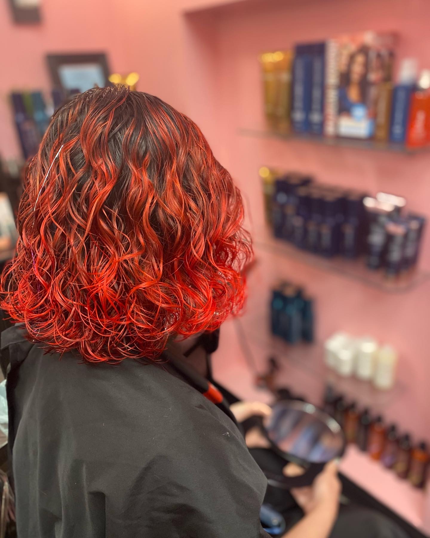 Person with curly, shoulder-length hair dyed red, facing shelves with hair products, in a salon or store setting.