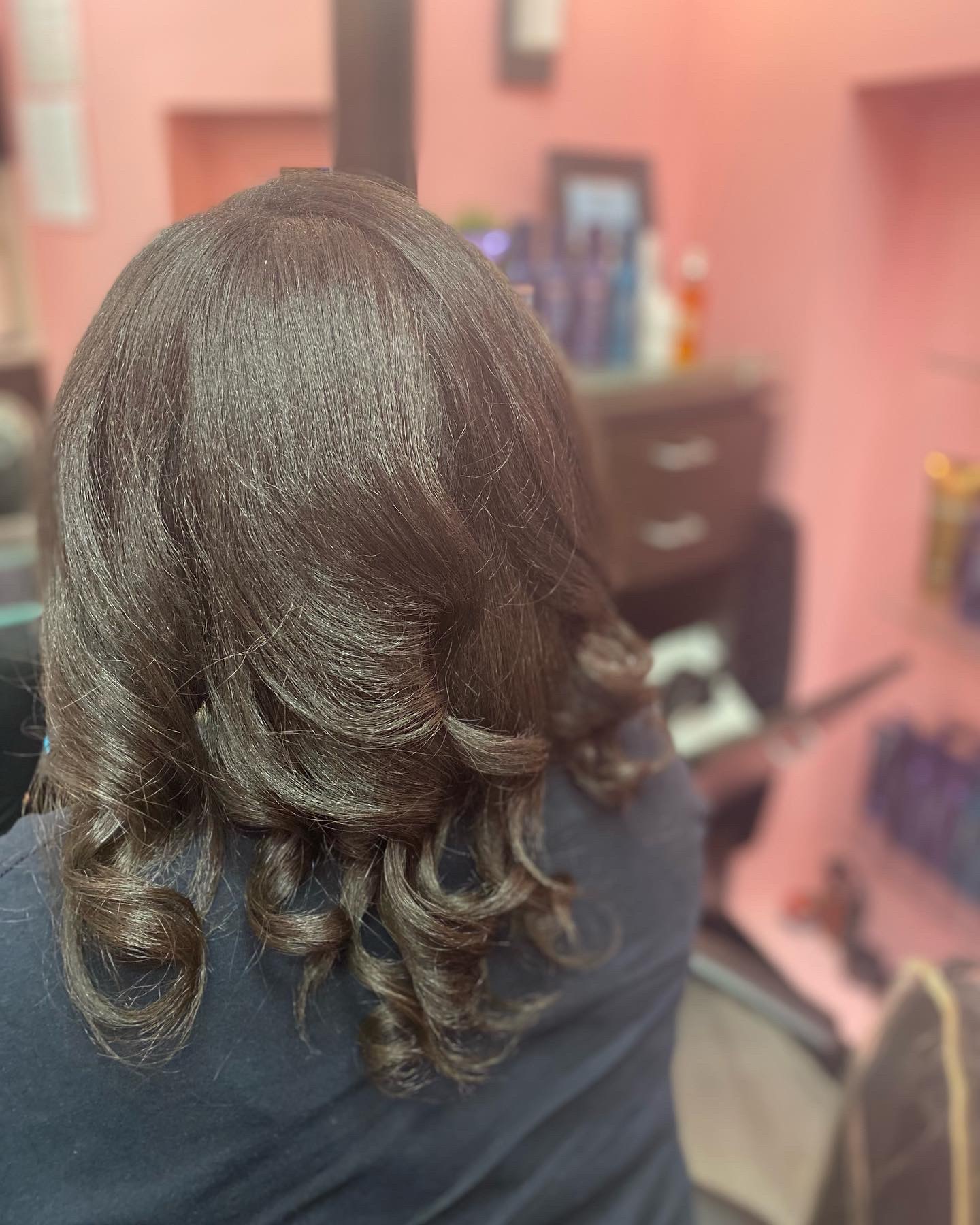 Back view of a person with long, dark, wavy hair in a salon, with pink walls and salon furniture visible.