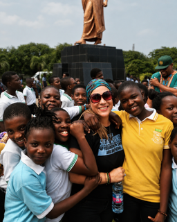 Group of children and a woman smiling in front of a large statue on a pedestal, outdoors with trees in background.
