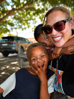 Woman wearing sunglasses smiling with two children outdoors, one child covering mouth, trees and parked cars in background.