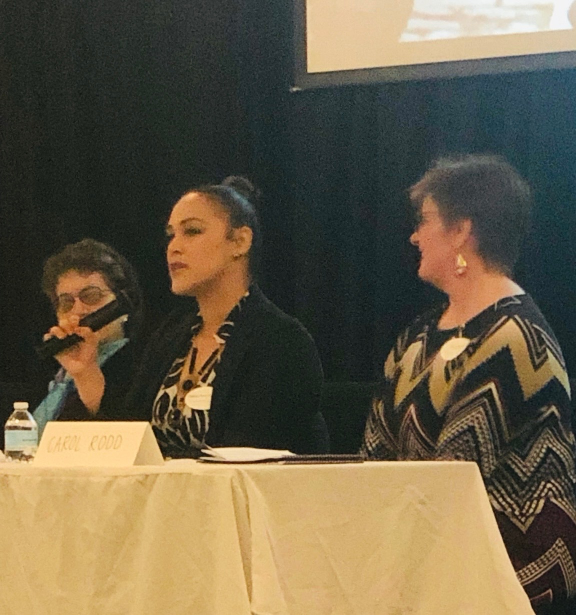 Three women seated at a table, one speaking into a microphone, with a black background and a screen above.