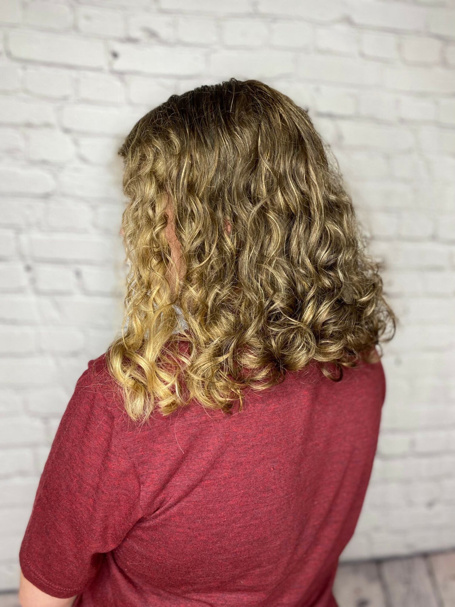 Person with curly blonde hair wearing a red top, facing away from the camera, standing in front of a white brick wall.