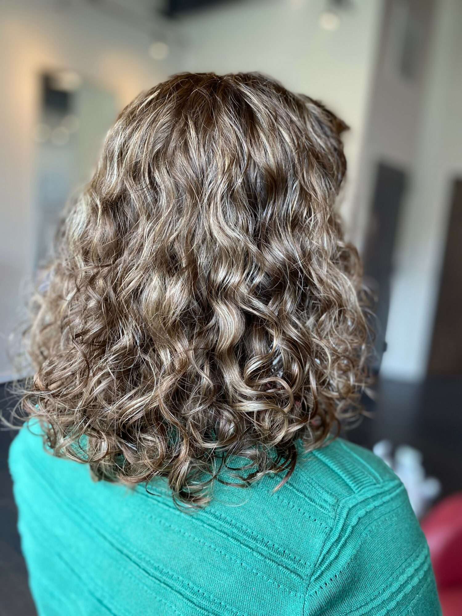 Back of a person with shoulder-length curly hair, wearing a teal top, in an indoor setting.