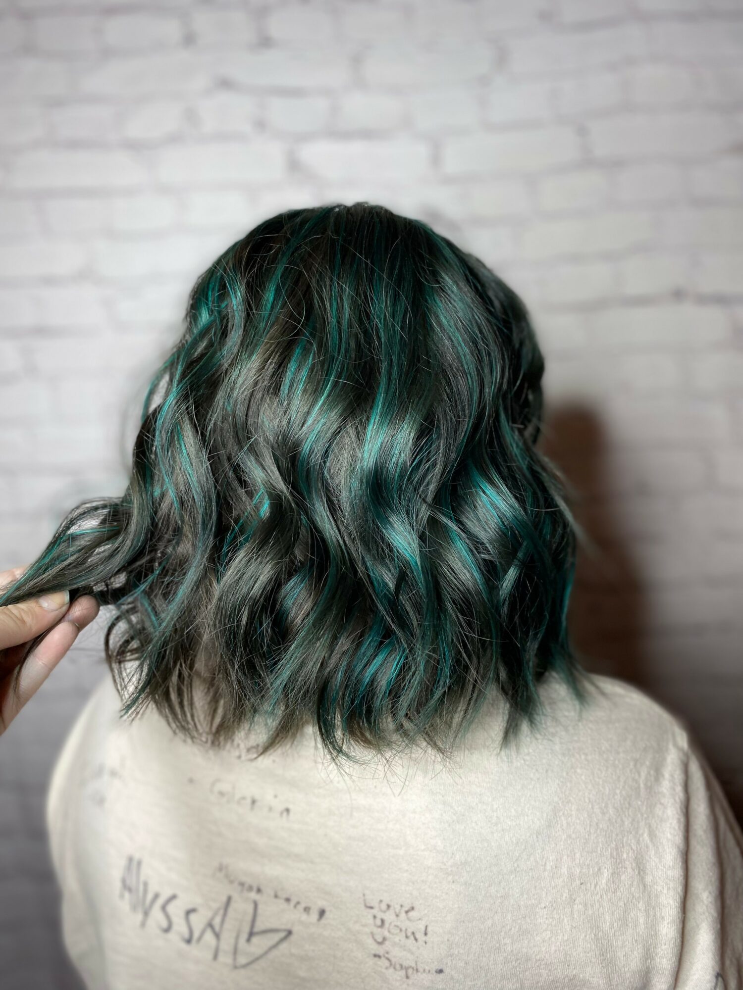 Back of a person's head with wavy, shoulder-length hair dyed in shades of black and teal, against a white brick wall.