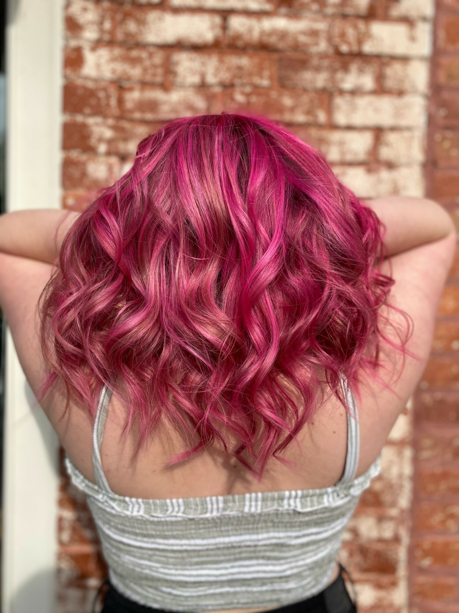 Back view of a person with shoulder-length, curly, pink hair, standing outdoors against a brick wall.