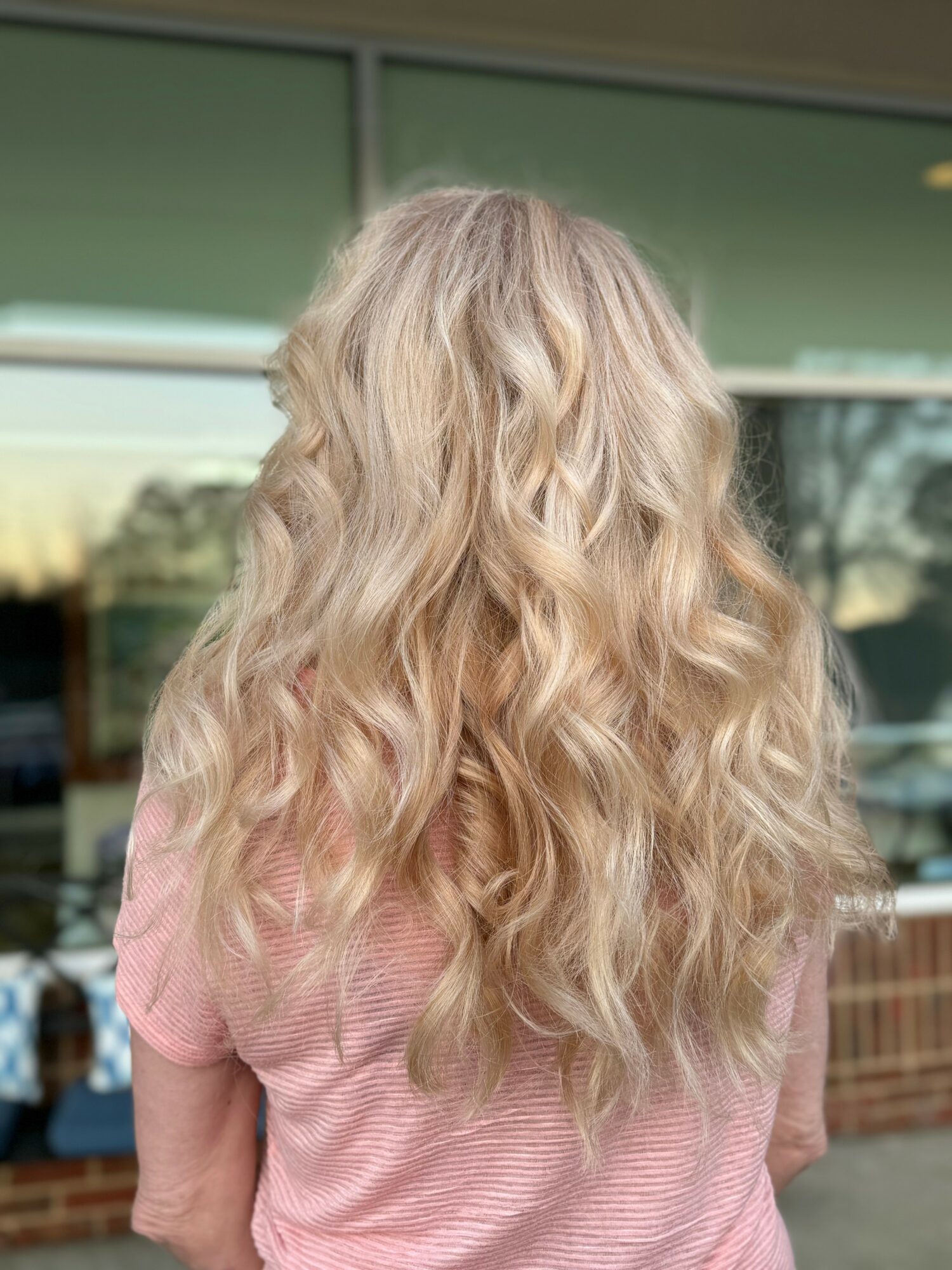 Back view of a person with long, wavy blonde hair wearing a pink top, standing outside a building with glass windows.