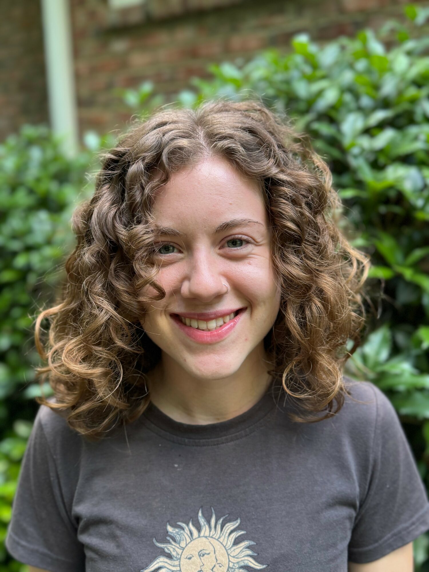 Young woman with curly hair smiling outdoors, greenery in background.
