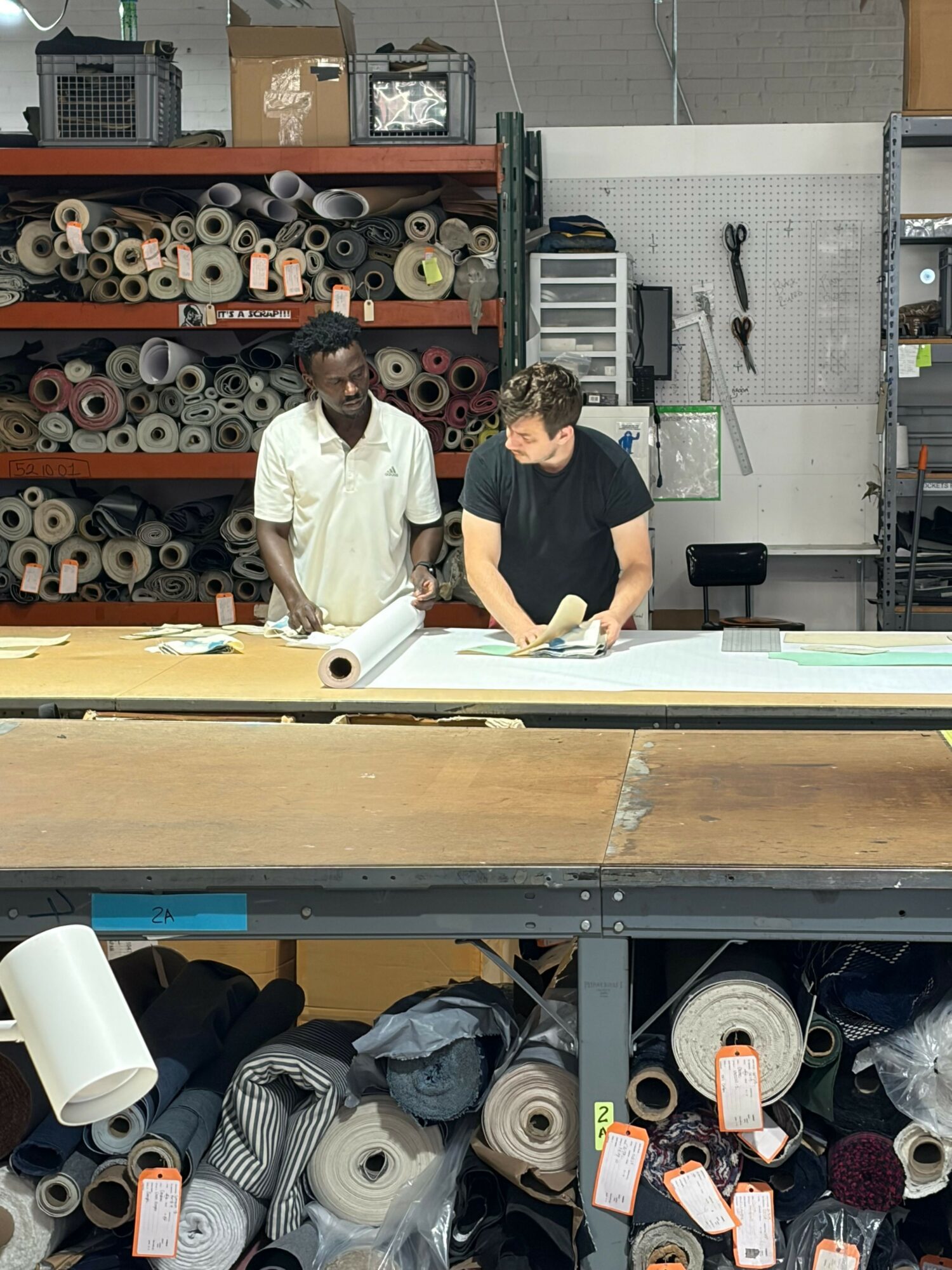 Two people working at a table with fabric rolls and sewing supplies in a workshop.