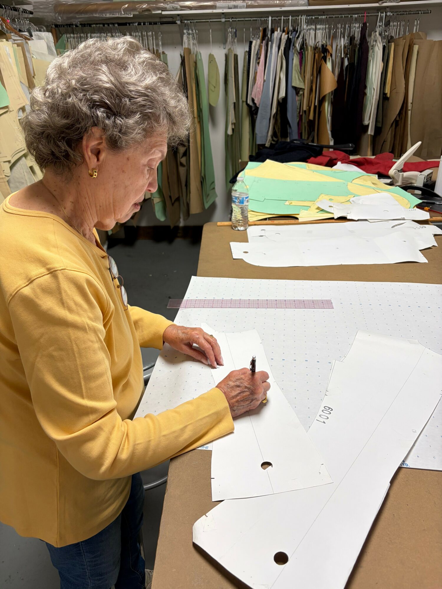 An elderly woman with curly gray hair wearing a yellow jacket writes on paper at a cluttered desk.