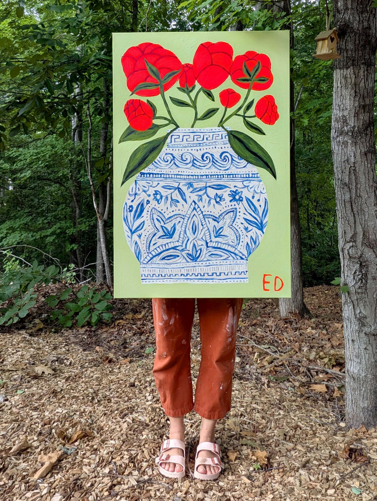 Person holding a large painting of a blue and white vase with red flowers outdoors, trees in background.