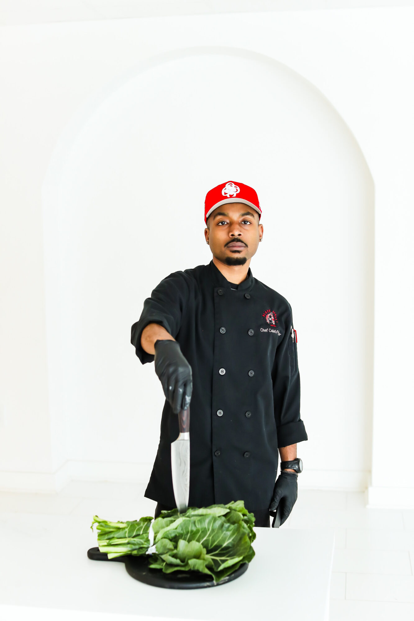 Person in black chef uniform and red cap stands behind a plate of leafy greens, holding a knife, in a white room.