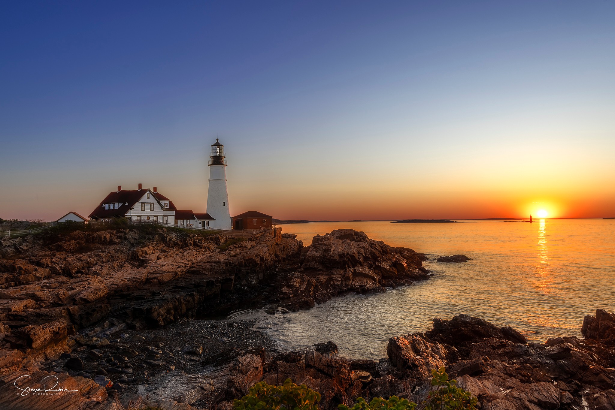 Lighthouse on rocky coast at sunset with sky transitioning from blue to orange, calm water reflecting the sunset, and a building nearby.