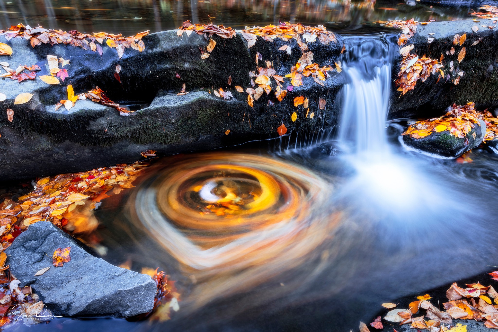 Stream with swirling water and fallen autumn leaves, small waterfall flowing over rocks, surrounded by colorful leaves on rocks and water.